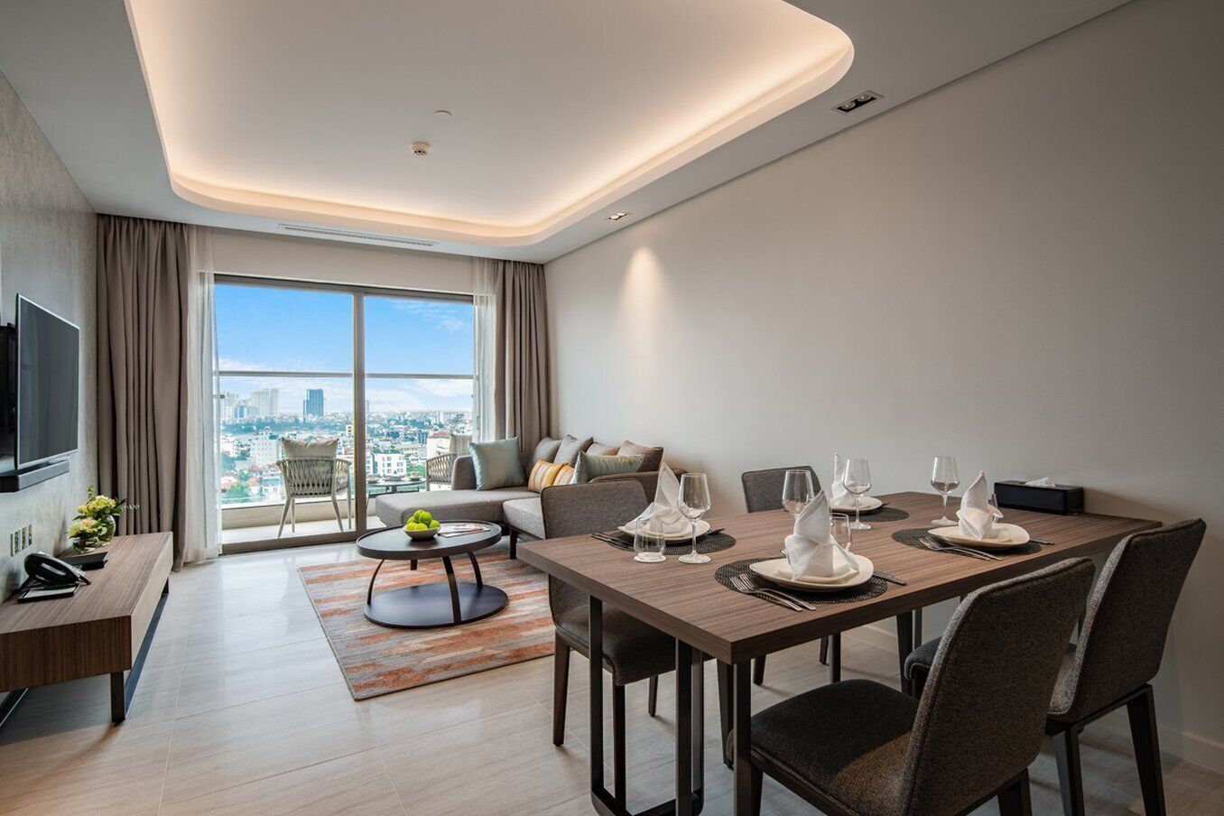 A modern hotel room with a dining area, a sofa, a TV, and a city view.
