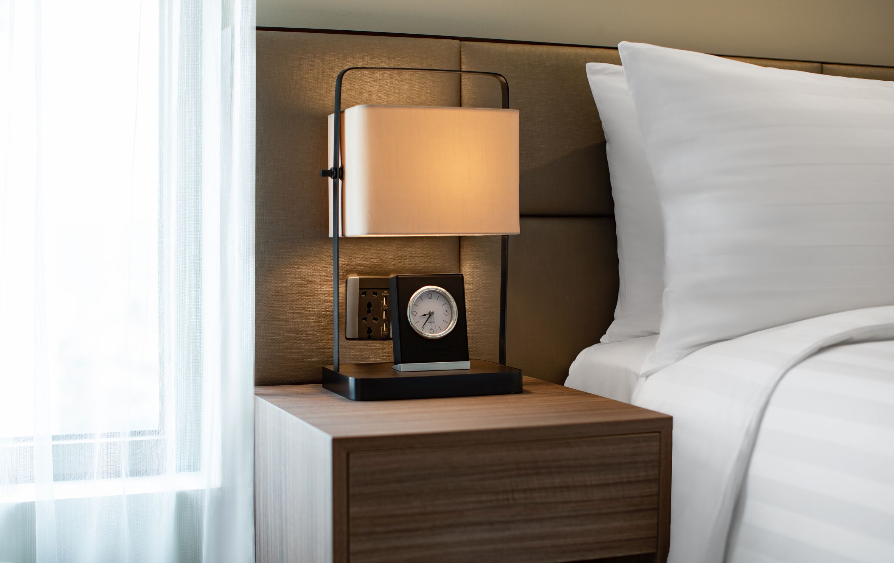 A neatly made bed with white linens, a bedside lamp, and a clock on the nightstand.