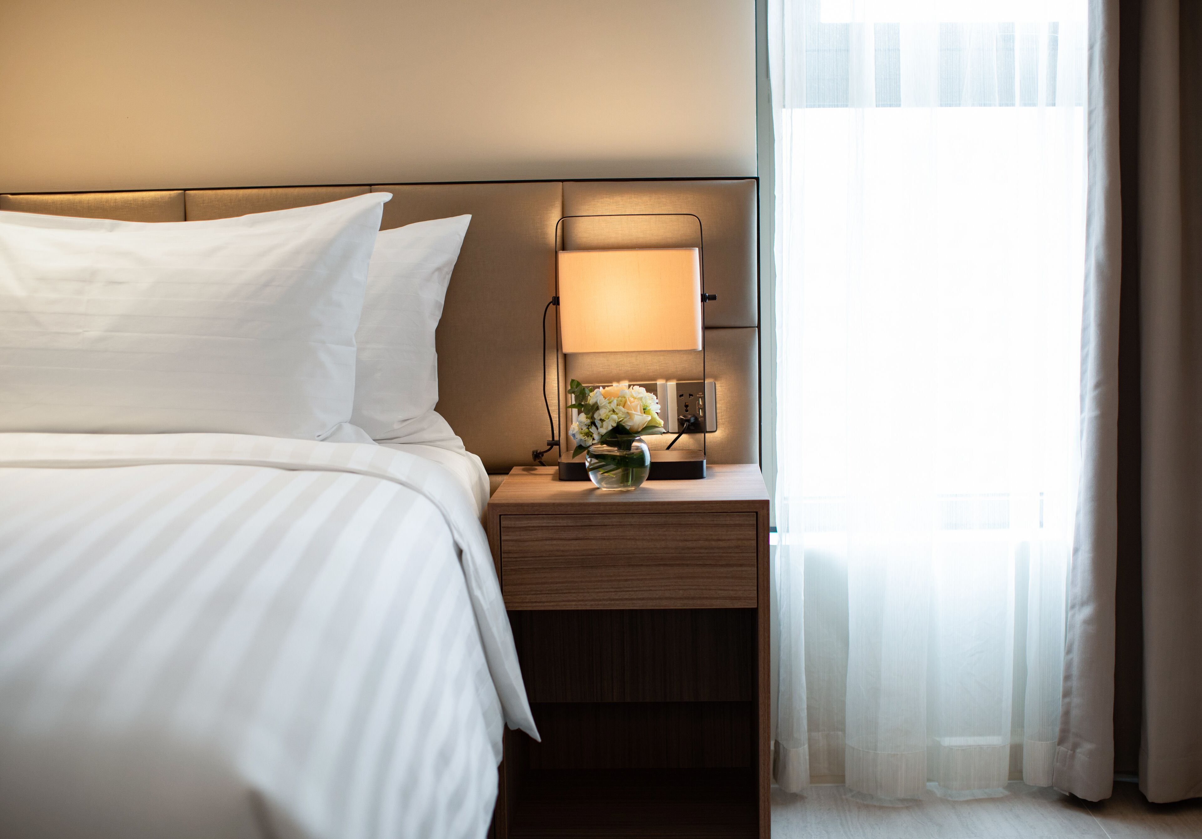 A hotel room with a neatly made bed, a bedside table with a lamp, and a window with sheer curtains.