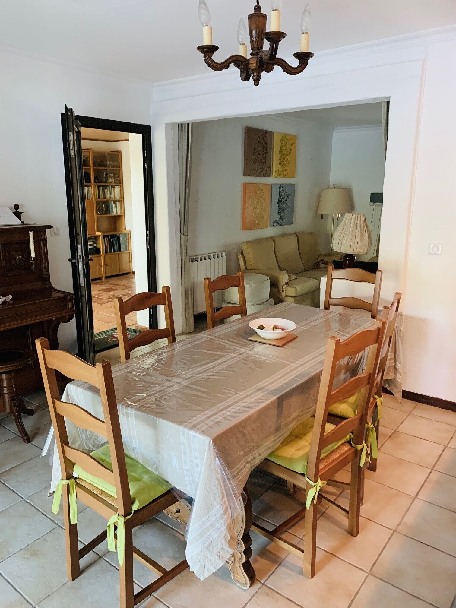 Un coin repas avec une table recouverte d’une protection, entourée de chaises en bois dotées de coussins verts. Un lustre est suspendu au-dessus de la table, et une bibliothèque est visible au fond.
