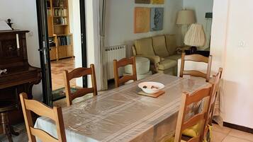 Un coin repas avec une table recouverte d’une protection, entourée de chaises en bois dotées de coussins verts. Un lustre est suspendu au-dessus de la table, et une bibliothèque est visible au fond.