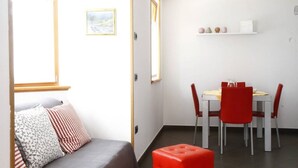 Una stanza piccola e luminosa con un letto, un tavolo da pranzo con sedie rosse e uno scaffale bianco con oggetti decorativi.