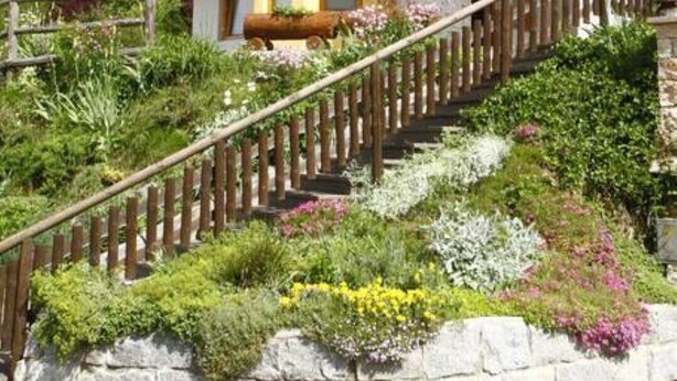 Un chalet a due piani con un balcone in legno, un muro di sostegno in pietra e una scala che conduce all'ingresso.
