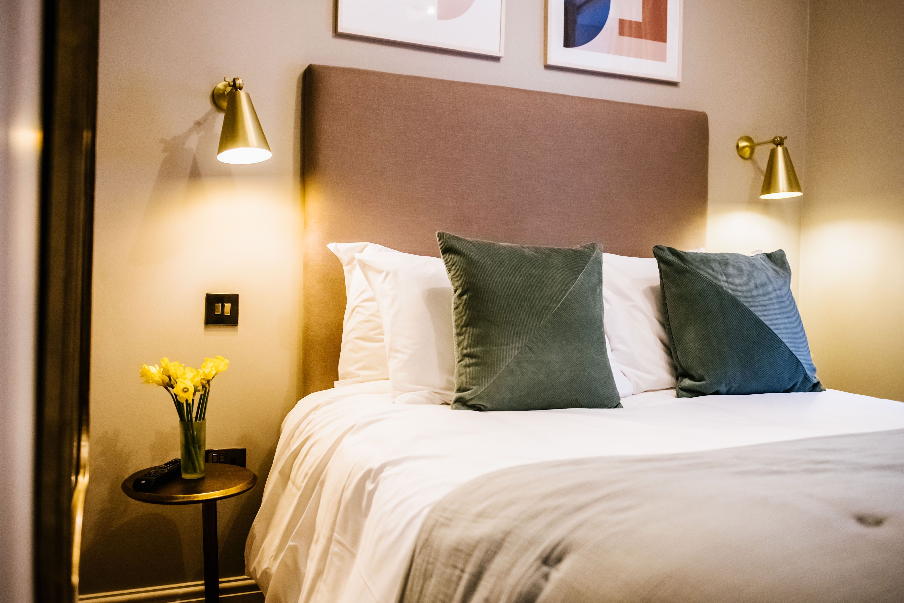 A neatly made bed with white and green pillows, a vase of yellow flowers, and two bedside lamps on a small table beside the bed.