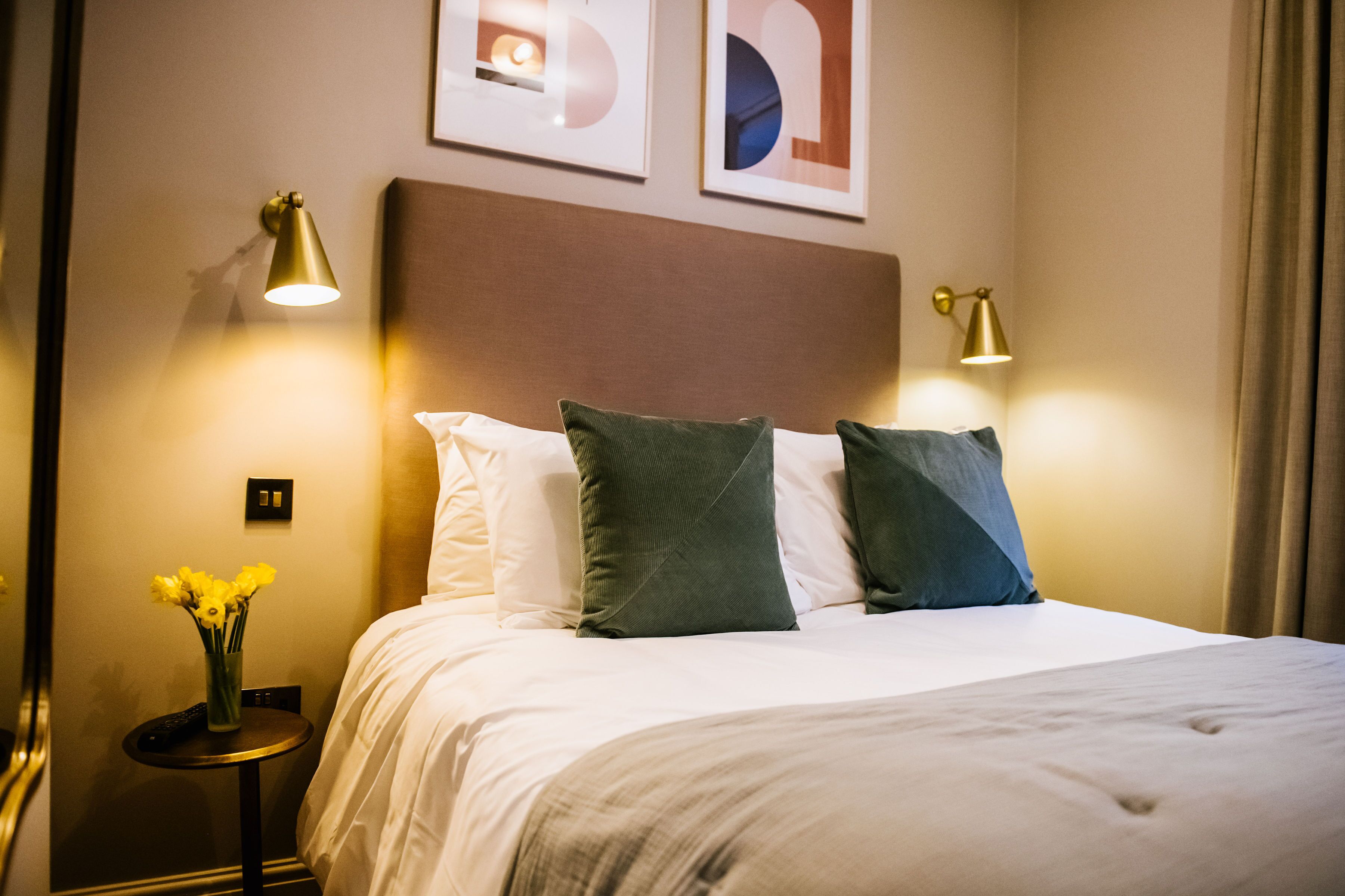 A hotel room with a neatly made bed, two bedside lamps, a small table with a vase of flowers, and framed artwork on the wall.