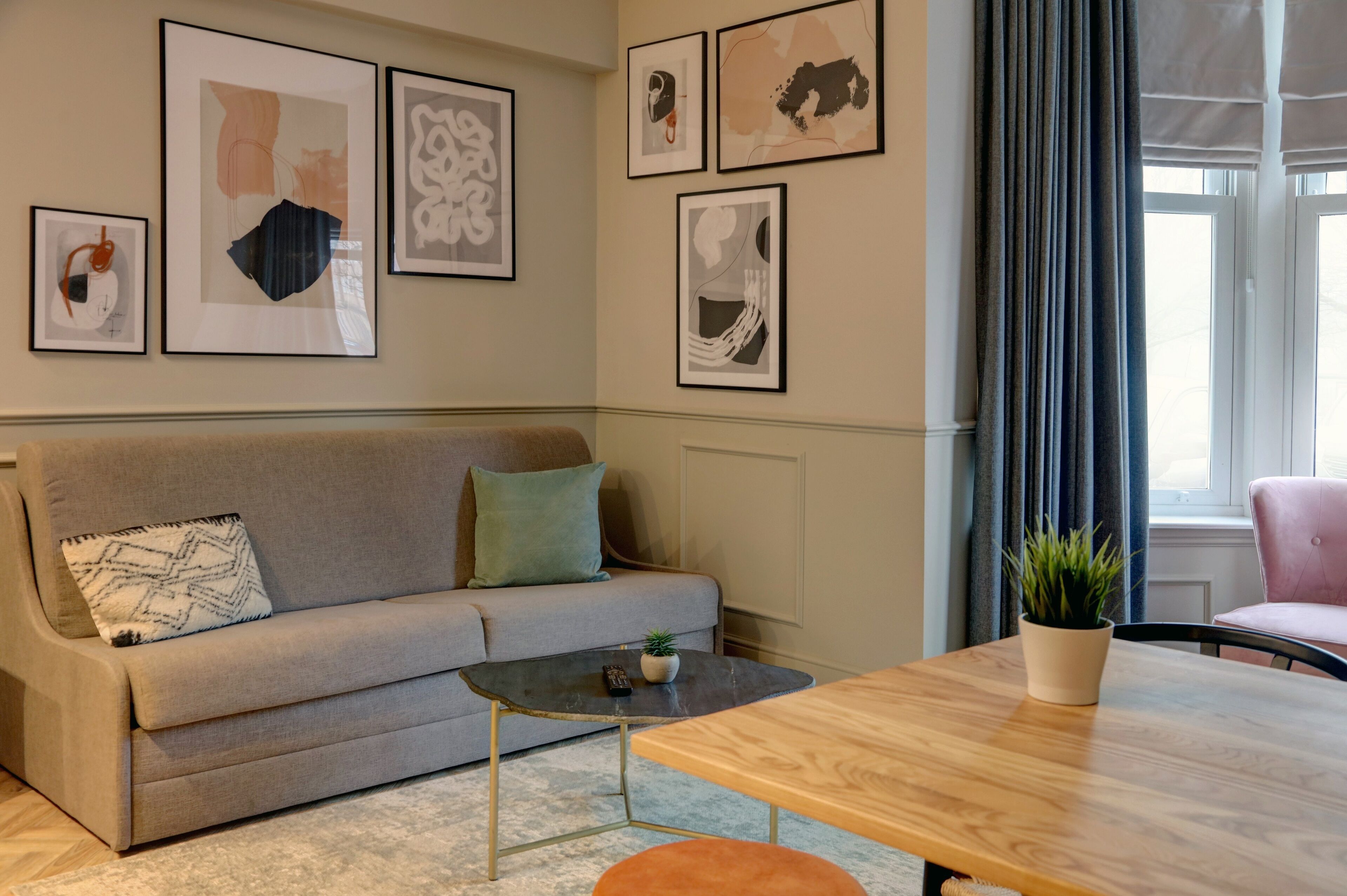 Un salon moderne avec un canapé gris, une petite table basse et une table à manger en bois. Des œuvres d’art encadrées ornent le mur, et une fenêtre est agrémentée de rideaux.