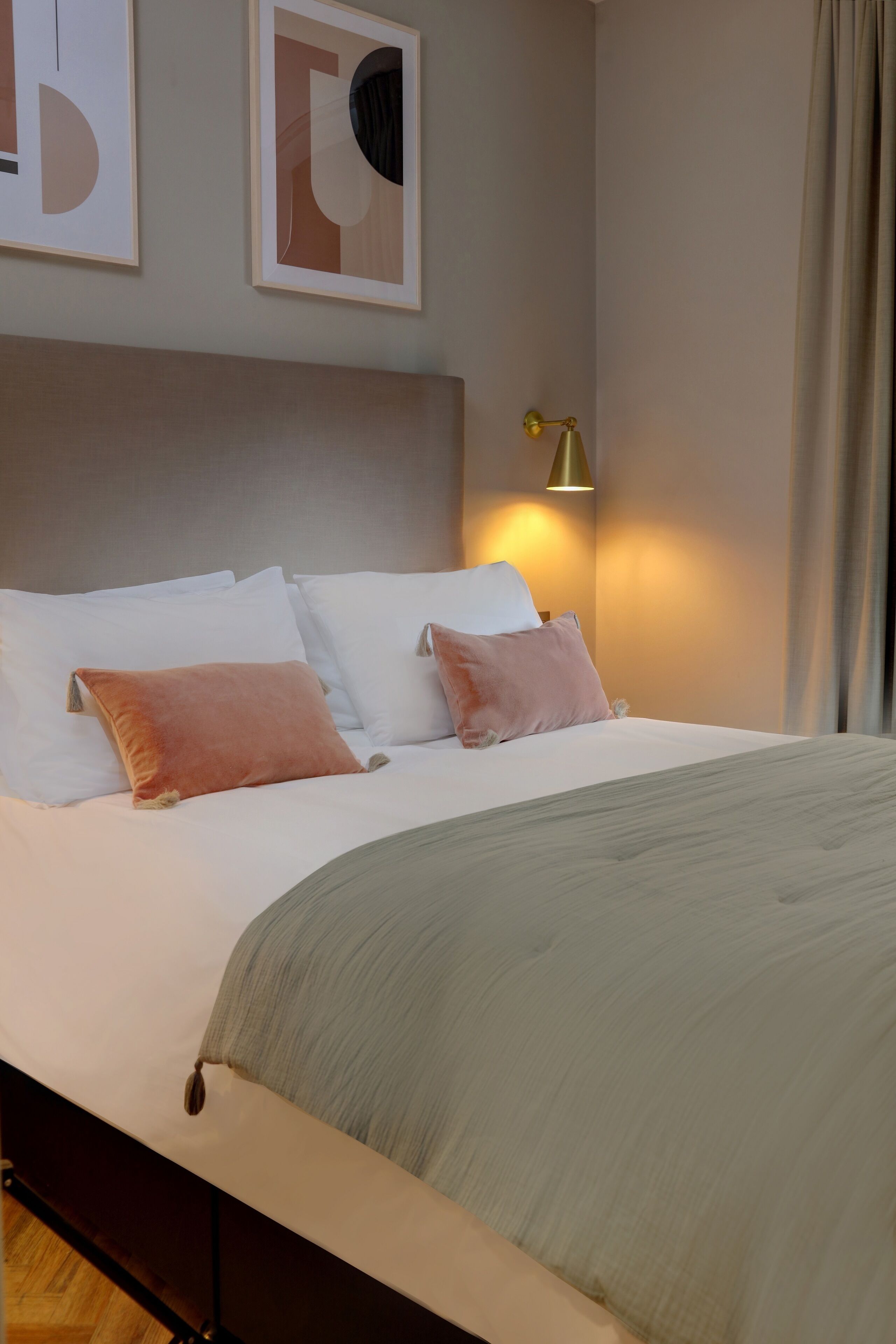 Una cama bien hecha con almohadas blancas y rosas, una manta gris y una lámpara de pared dorada.