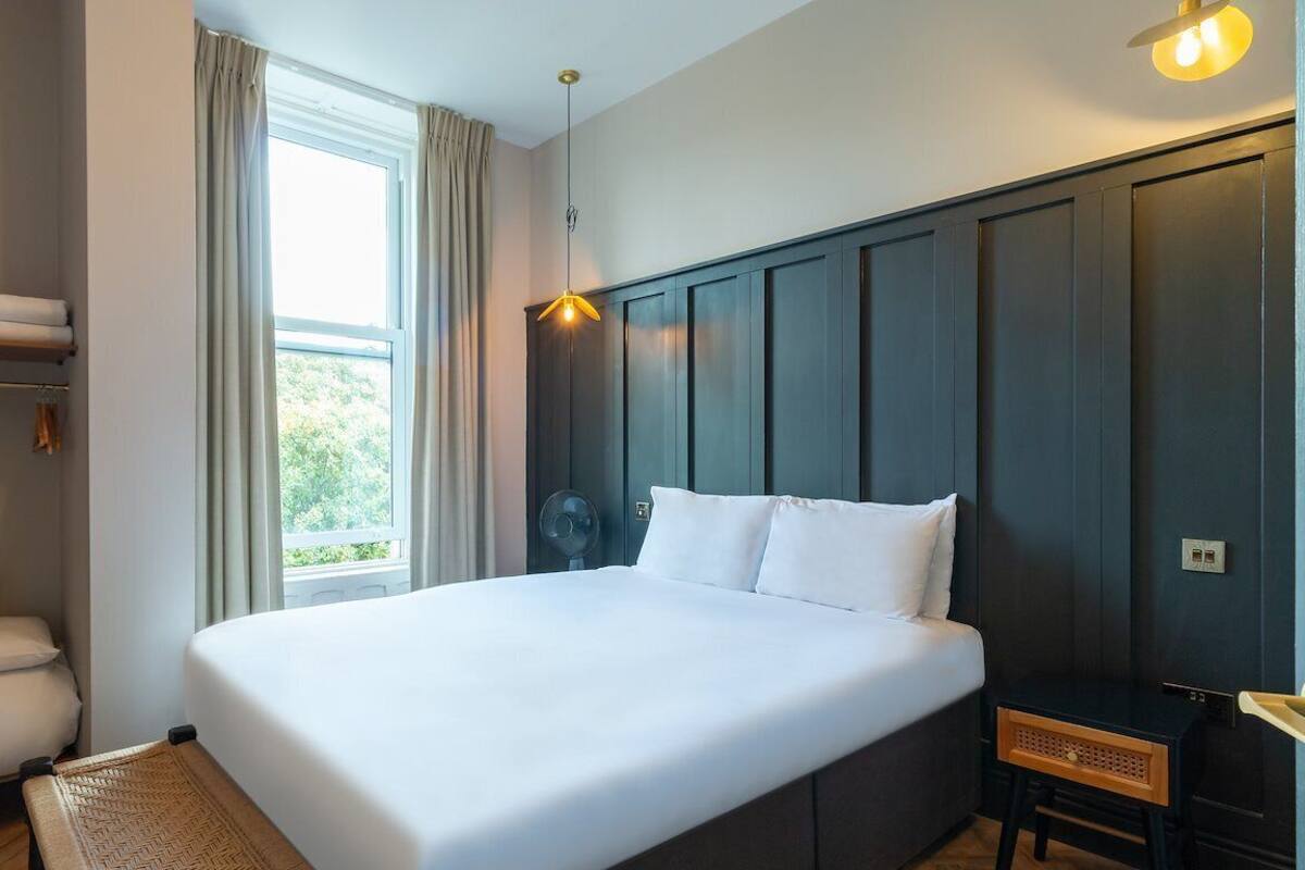 A neatly arranged bedroom with a bed, a bedside table, a shelf with towels, and a window with curtains.
