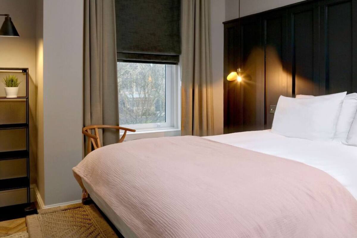A bedroom with a large bed, a nightstand, a lamp, a window with curtains, and a view of trees outside.