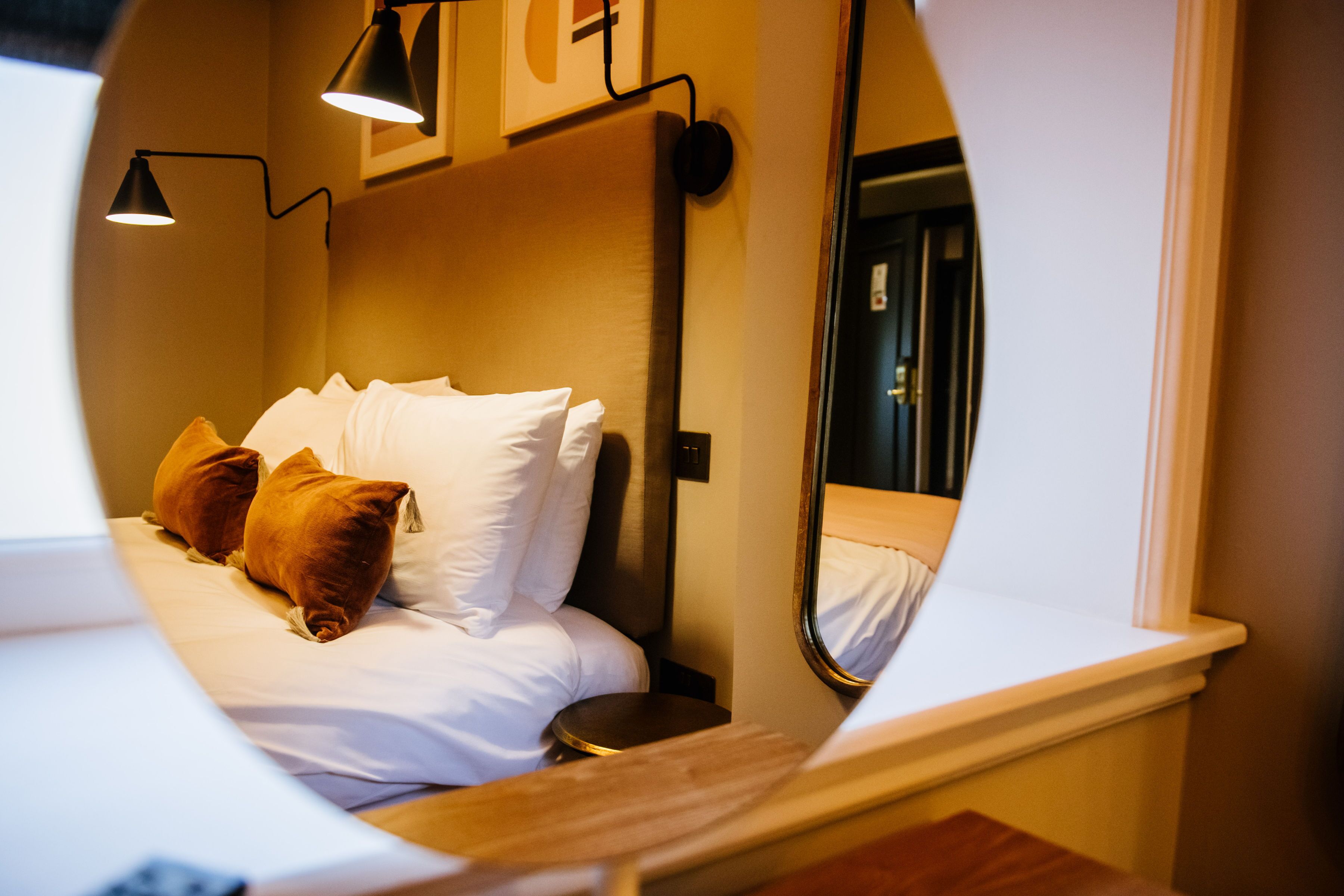 A circular view of a hotel room with a bed, pillows, a nightstand, and a mirror reflecting the room.