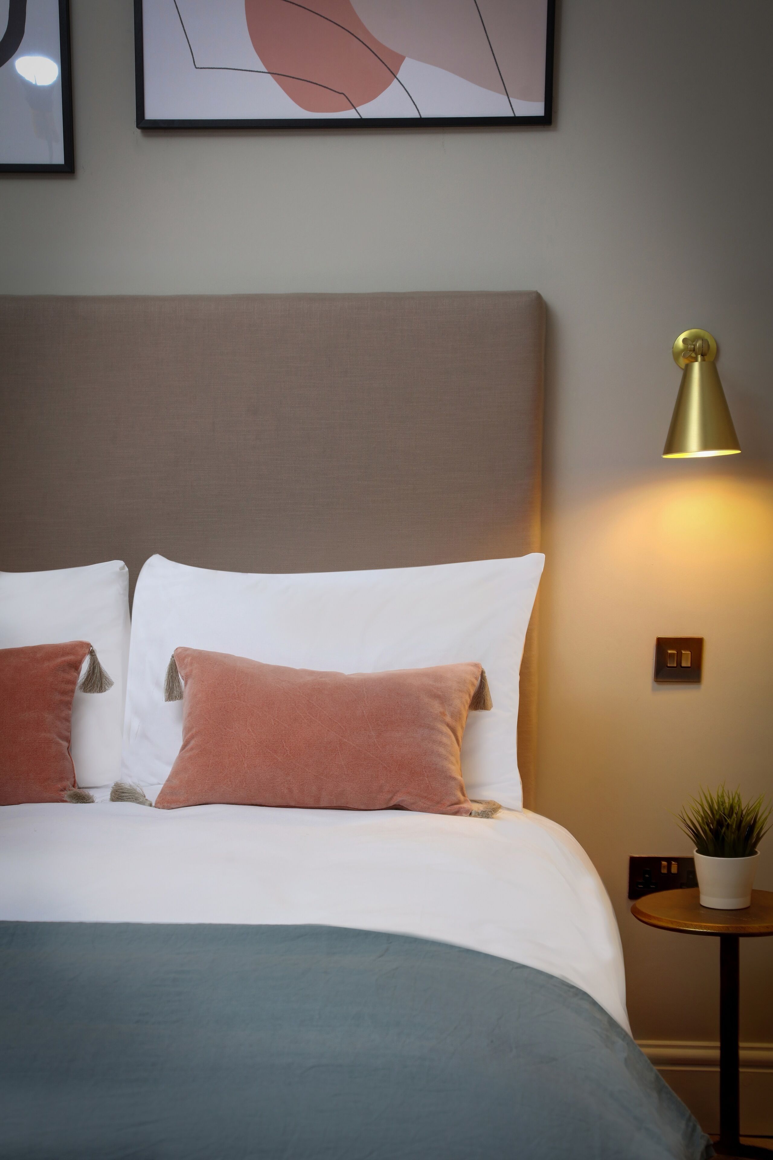 A neatly made bed with white and pink pillows, a green throw blanket, and a small round table with a plant beside it.