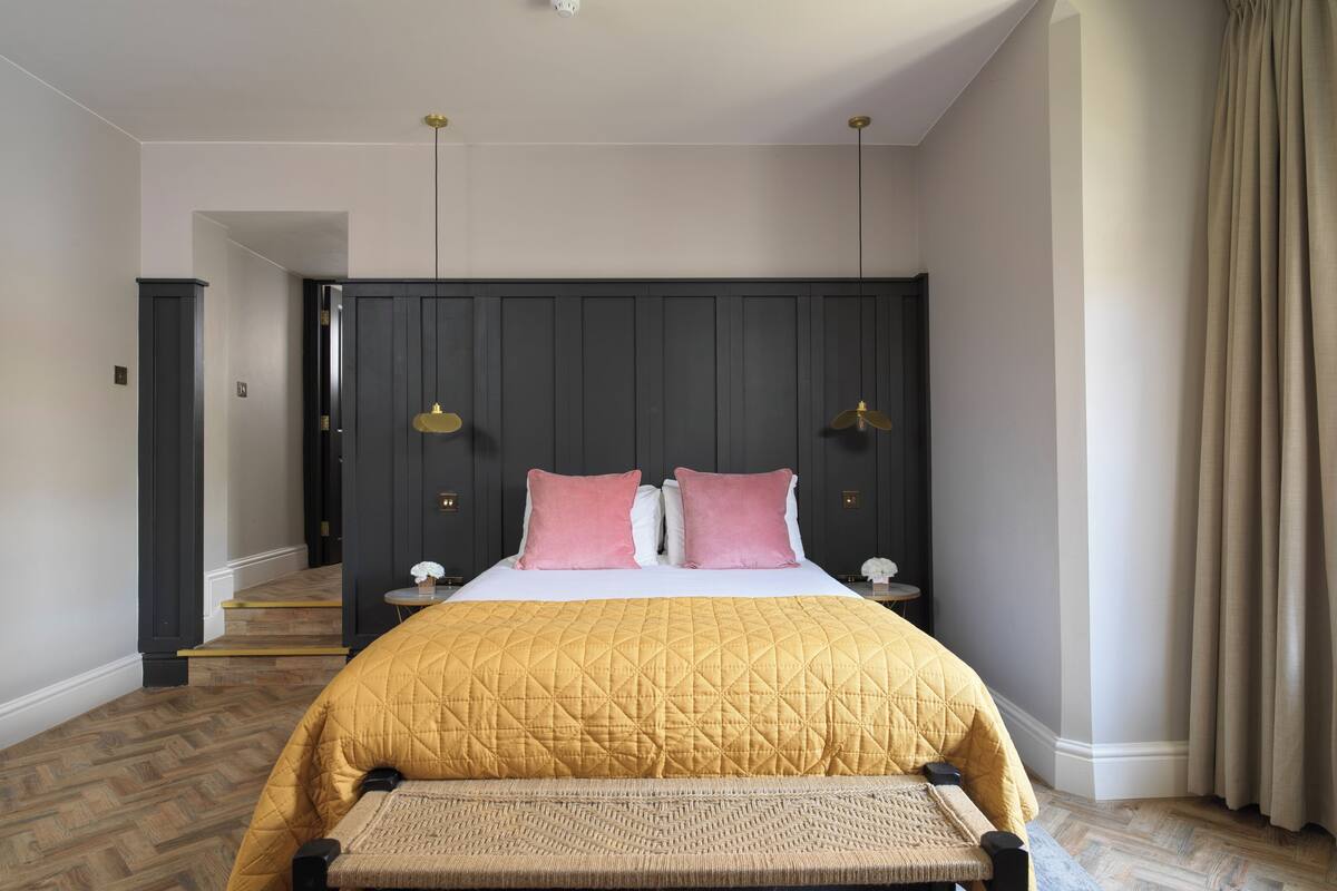 A bedroom with a large bed, a bench, a dark headboard, two bedside lamps, and a staircase in the background.