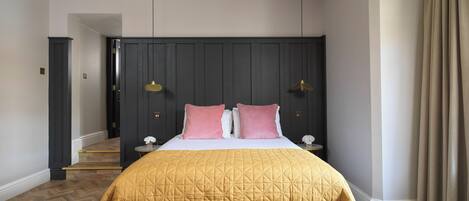 A bedroom with a large bed, a bench, a dark headboard, two bedside lamps, and a staircase in the background.