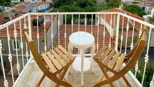 Un balcón con una mesa y dos sillas de madera, con vista a un pueblo de tejados rojos y cielo despejado.