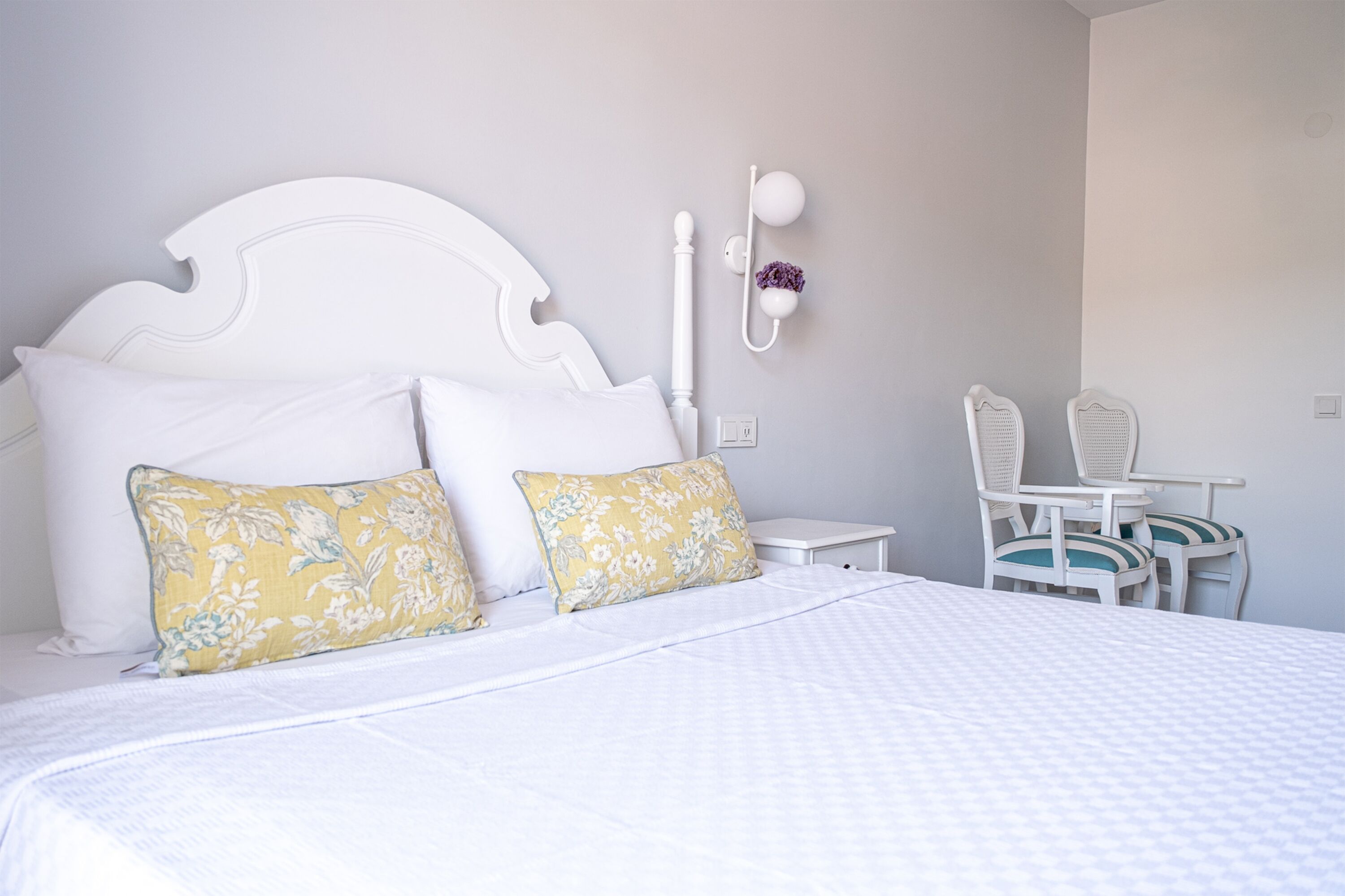 A neatly made bed with white bedding and two floral pillows, a white headboard, a small bedside table with a lamp, and a chair with a cushion.