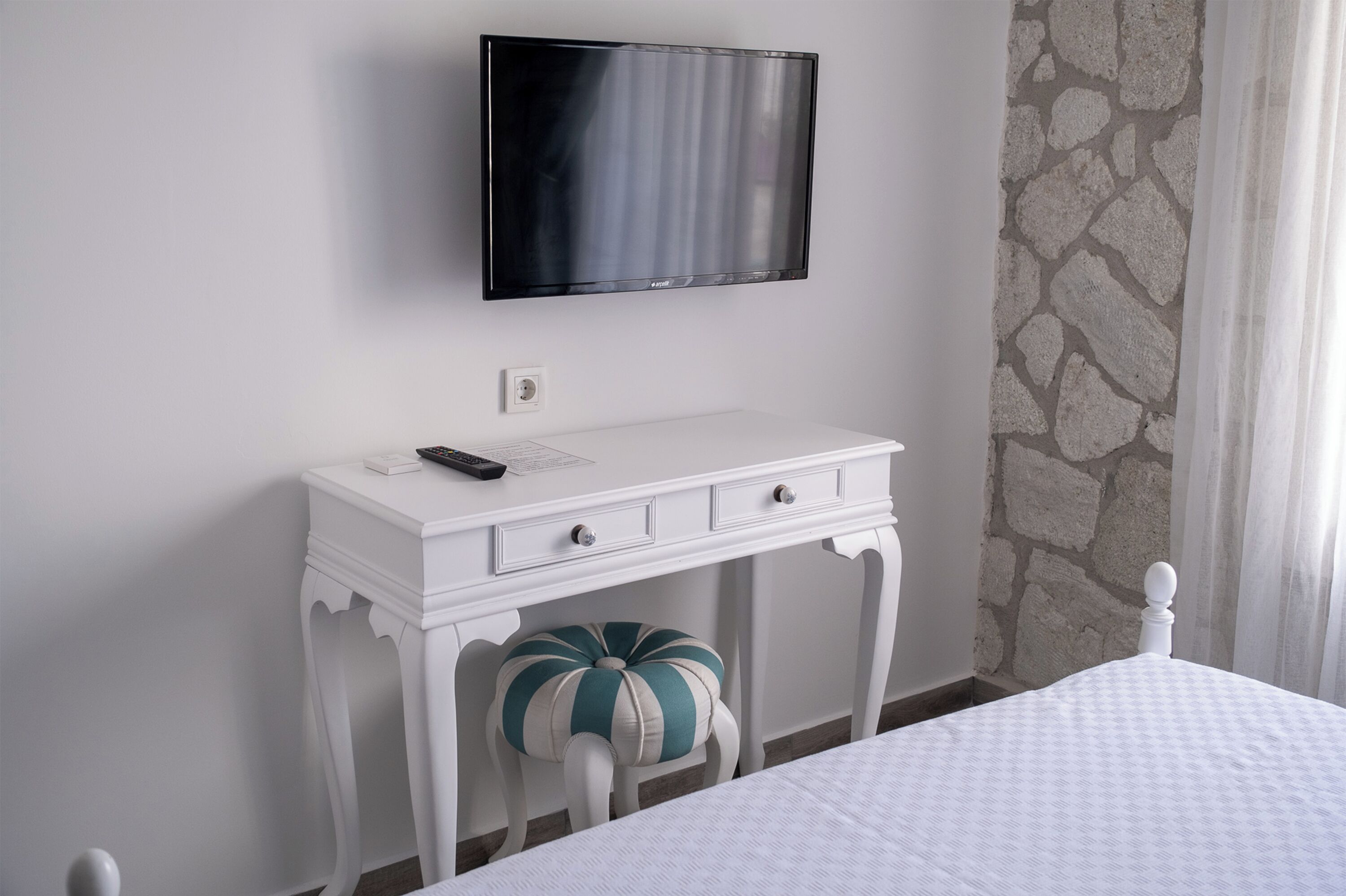 A neatly arranged bedroom with a bed, a white dresser with a remote control, a television mounted on the wall, and a stone-patterned wall.
