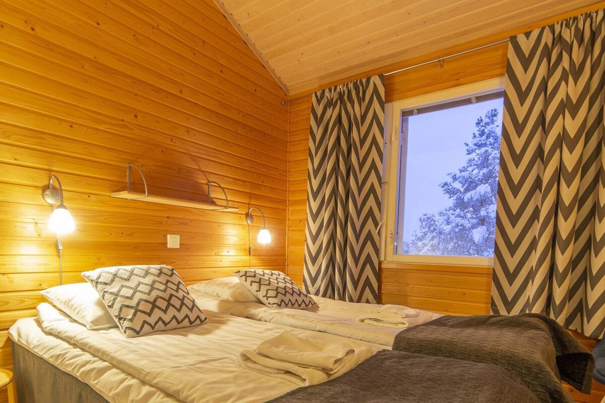 Un dormitorio de cabaña de madera con una cama, dos lámparas fijadas a la pared, una ventana con cortinas y un estante sobre la cama.