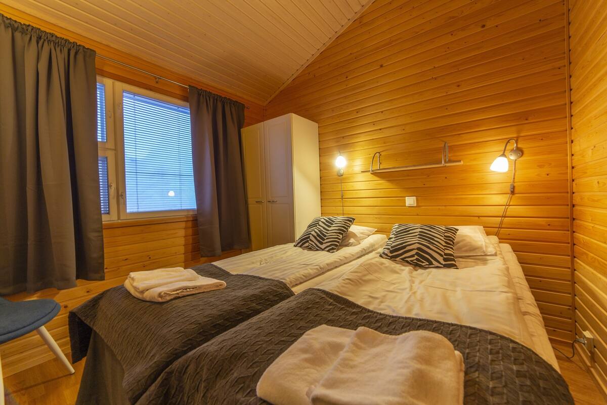 Una habitación con paredes de madera, dos camas con almohadas, un banco, una ventana con cortinas y una lámpara fijada a la pared.