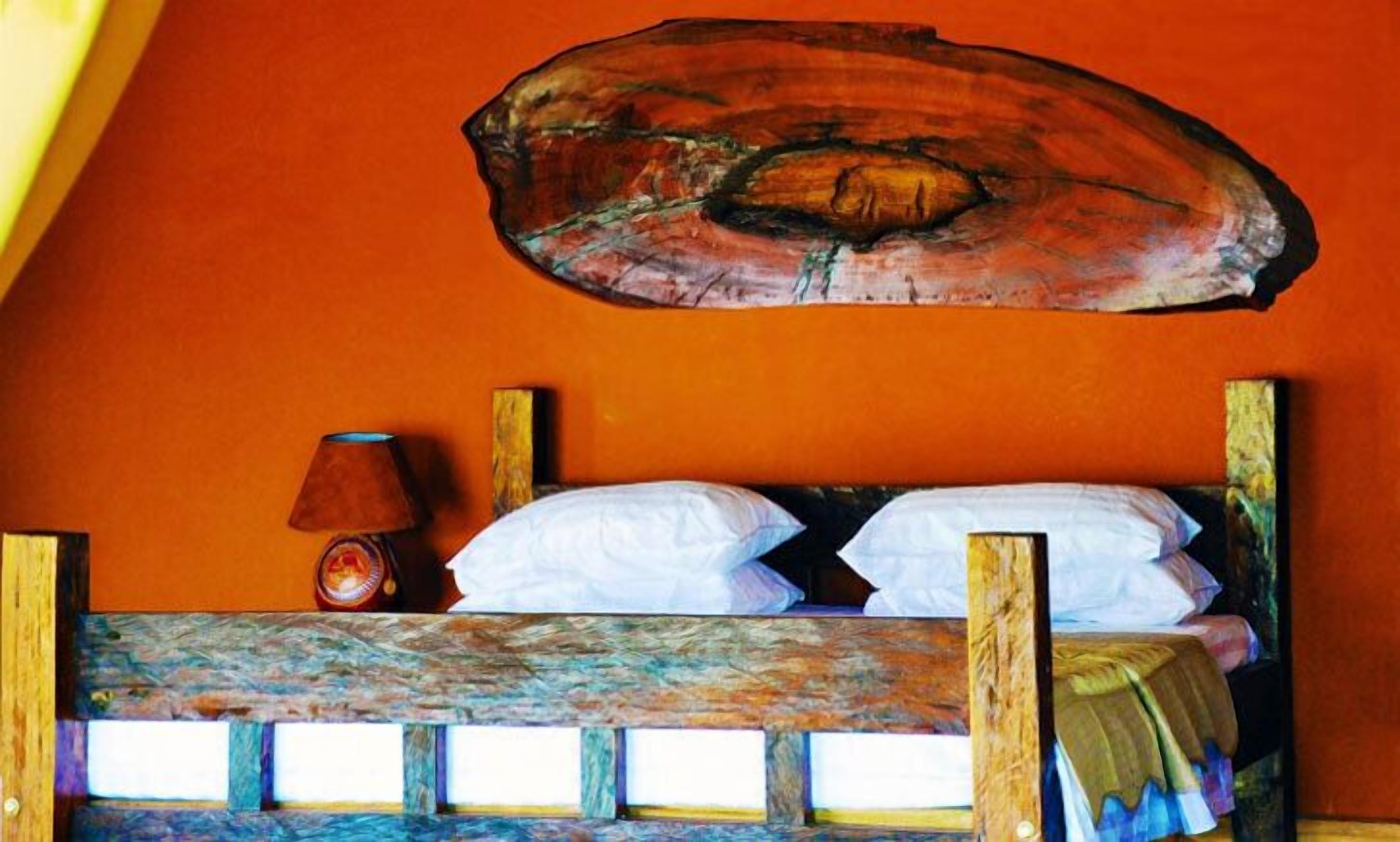 A bed with white pillows and a wooden headboard, a lamp, and a large abstract painting on an orange wall.