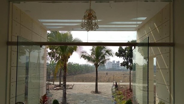 A view from inside a building with a glass wall, looking out to a landscaped area with palm trees and a red carpet leading to a door. - Somudra Bari Resort (Kalapara)
