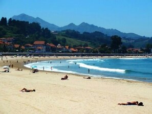 Ein Strand mit badenden und sonnenden Menschen, umgeben von Häusern und Bergen im Hintergrund.