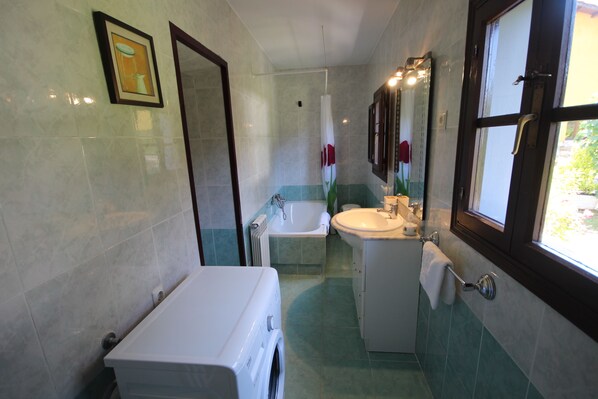 Ein Badezimmer mit Waschmaschine, Badewanne, Waschbecken und einem Fenster mit Blick ins Grüne.