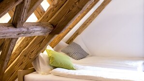 Un dormitorio tipo loft con techo de vigas de madera, cama con ropa blanca y sillón gris.