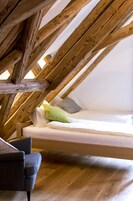 Quarto em estilo loft com teto de vigas de madeira, cama com roupa de cama branca e poltrona cinza.