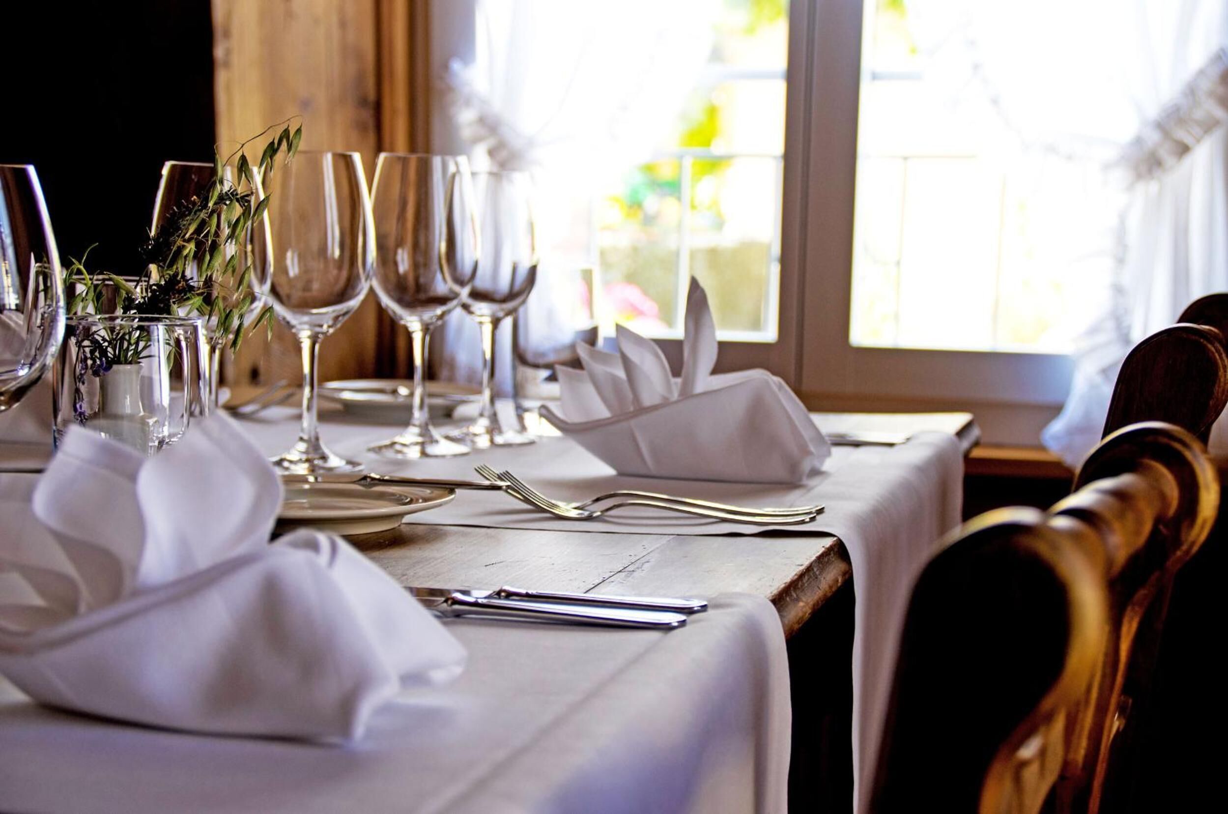 Una mesa de comedor dispuesta con manteles blancos, cubiertos y copas de vino, con una ventana al fondo.