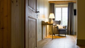 Una habitación con un armario de madera, una mesa con una lámpara, una silla y una ventana con cortinas.