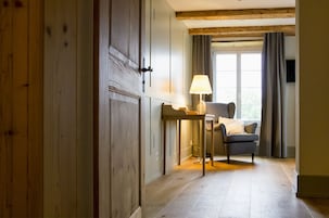 Quarto com guarda-roupa de madeira, uma mesa com abajur, uma cadeira e janela com cortinas.