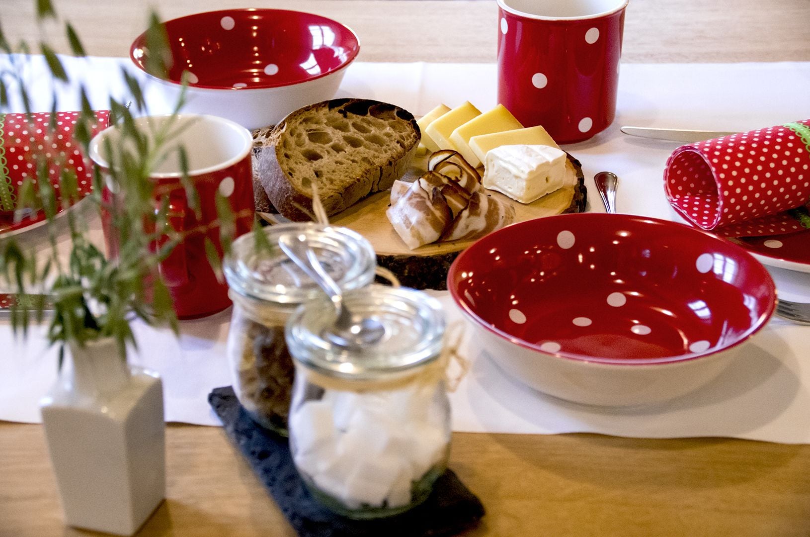 A table set with red and white polka dot dishes, jars of food, and a variety of cheeses and bread.