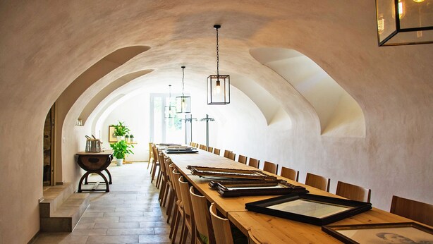 Una larga mesa de comedor con sillas, cuadros enmarcados y luces colgantes en una habitación con techos abovedados.