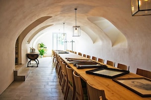 Uma longa mesa de jantar com cadeiras, quadros e luminárias pendentes num espaço com tetos abobadados.