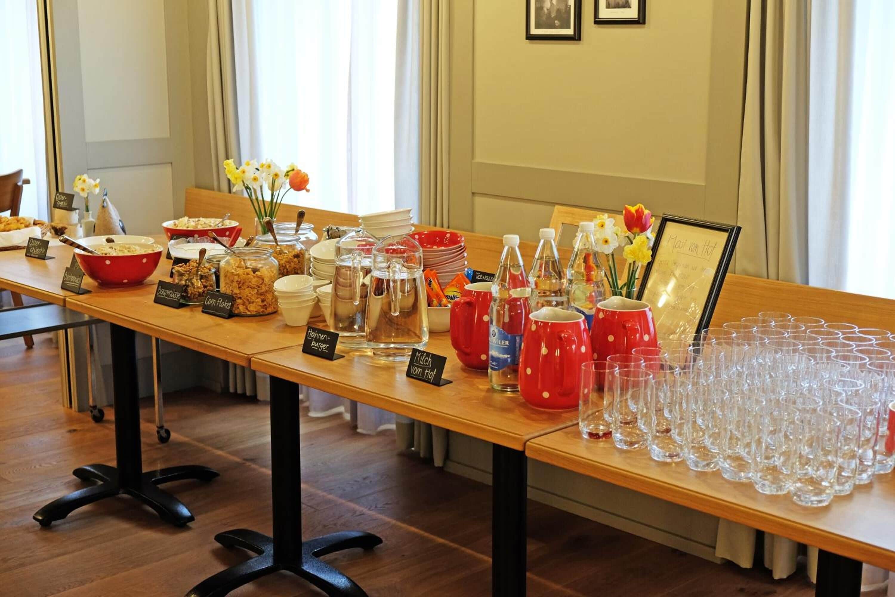 A buffet table with various food items and drink dispensers, arranged on a wooden table with a glassware set at the end.