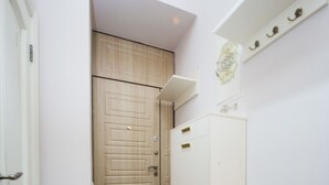 Un pasillo estrecho con una puerta de madera, un armario blanco con tiradores dorados y un pequeño estante con adornos.