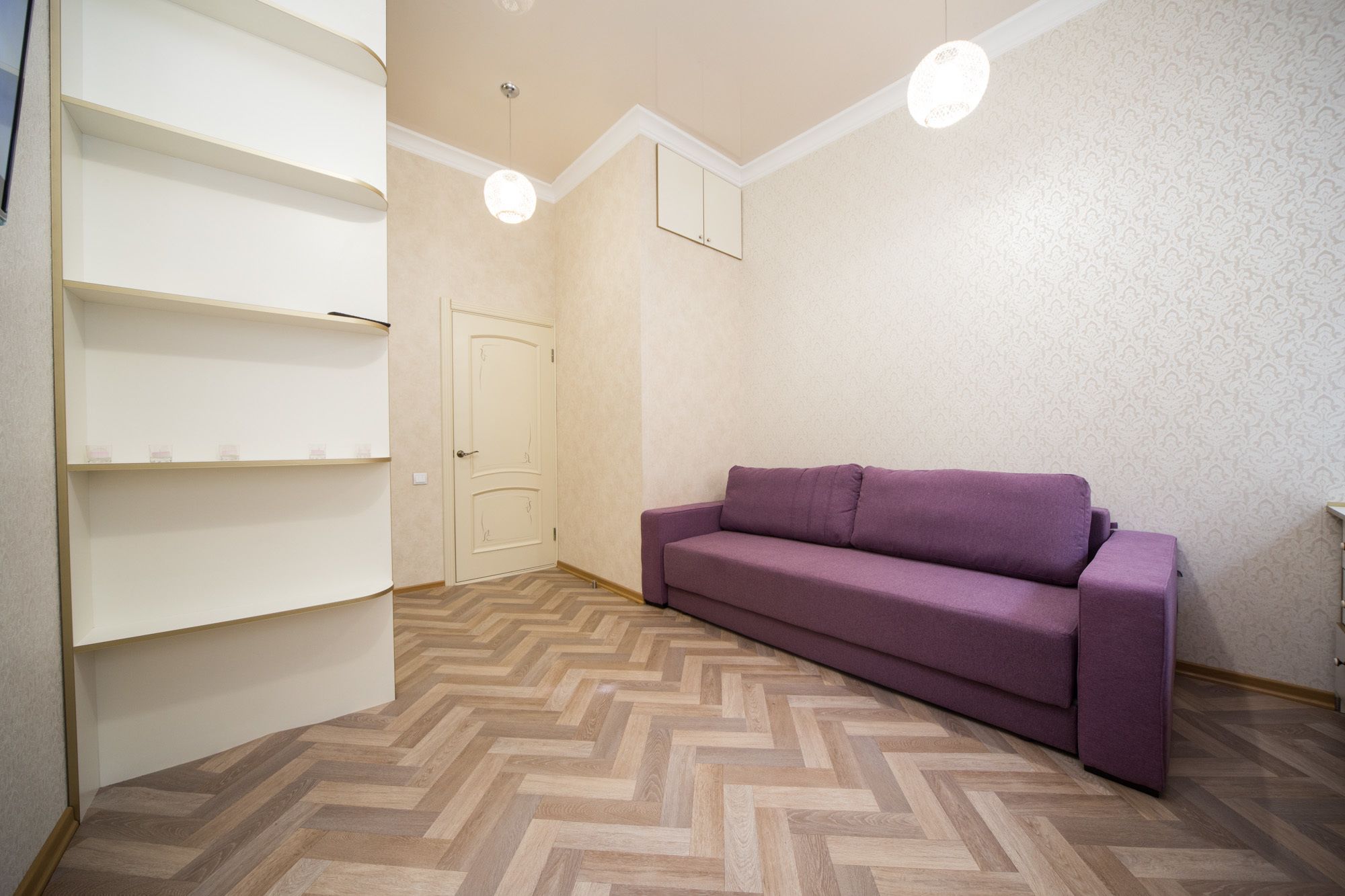 A room with a herringbone-patterned wooden floor, a purple sofa, and white shelving units.