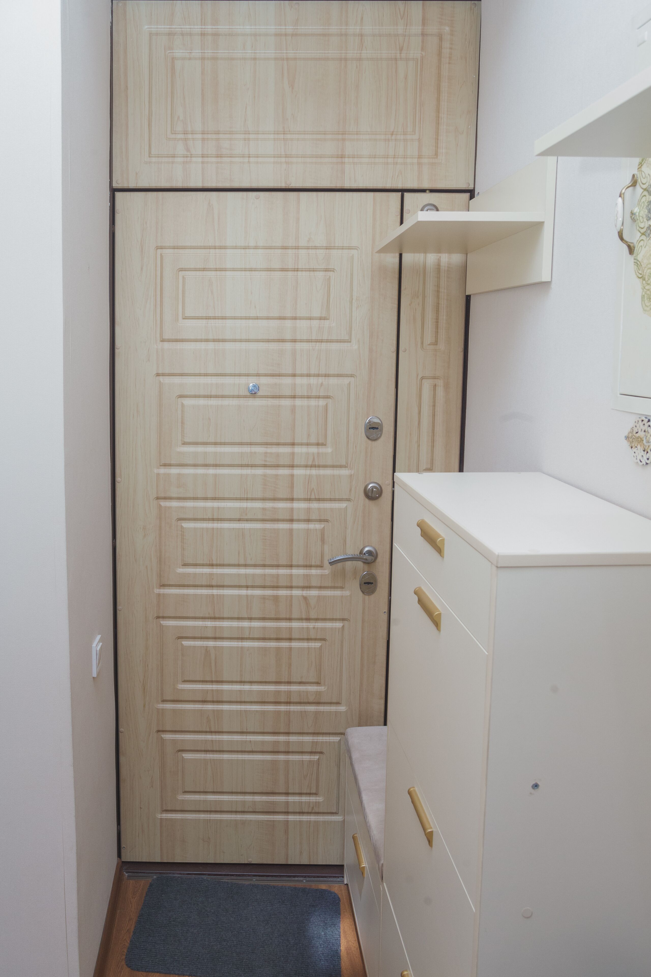 Una habitación pequeña con una puerta de madera, un armario blanco con cajones y un estante pequeño encima.
