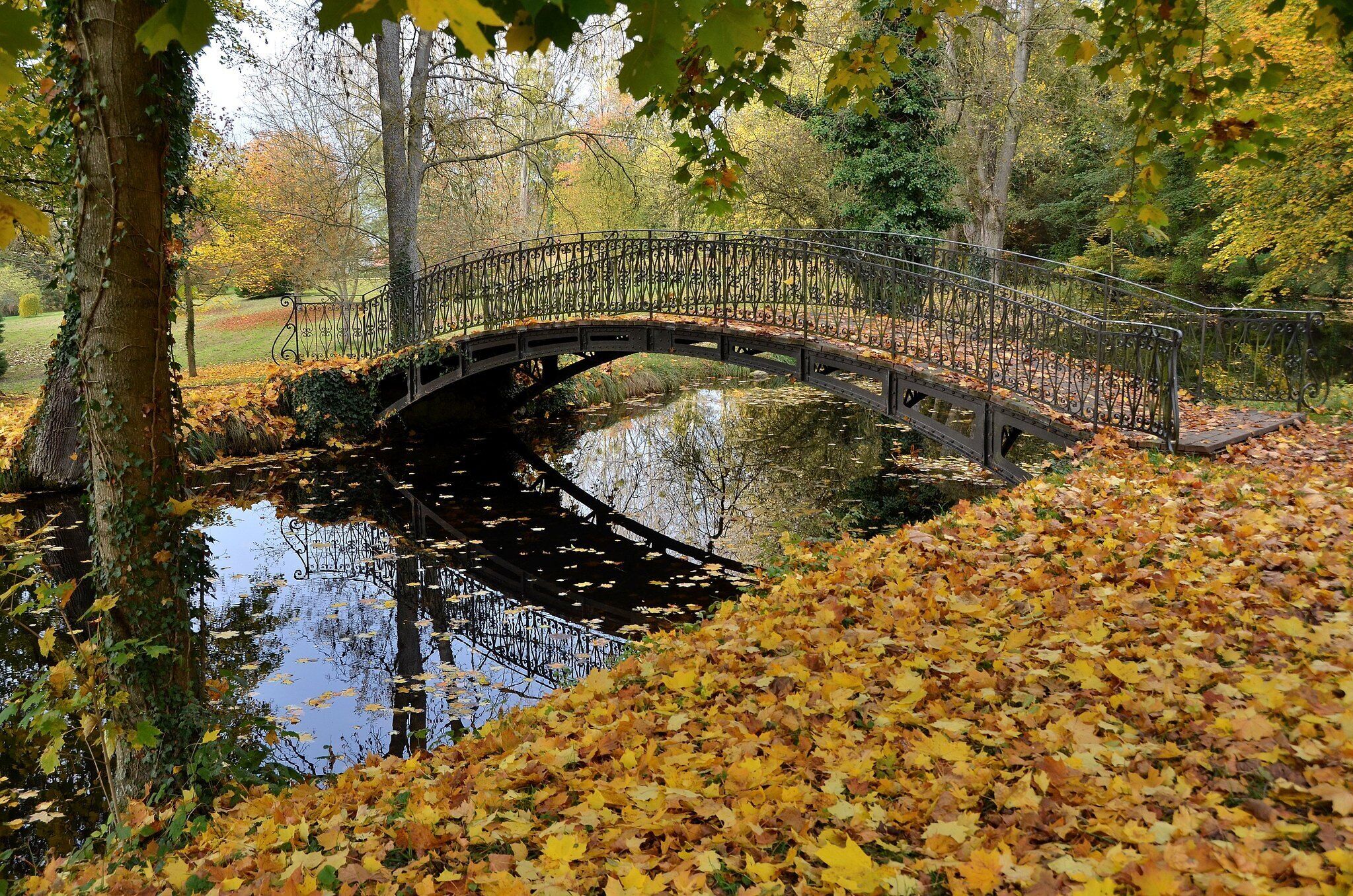A stone bridge over a calm water body surrounded by trees with autumn leaves.