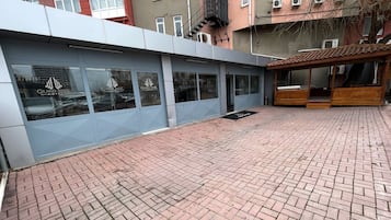 Exterior de hotel com pátio de tijolos, grandes janelas e área de estar coberta.