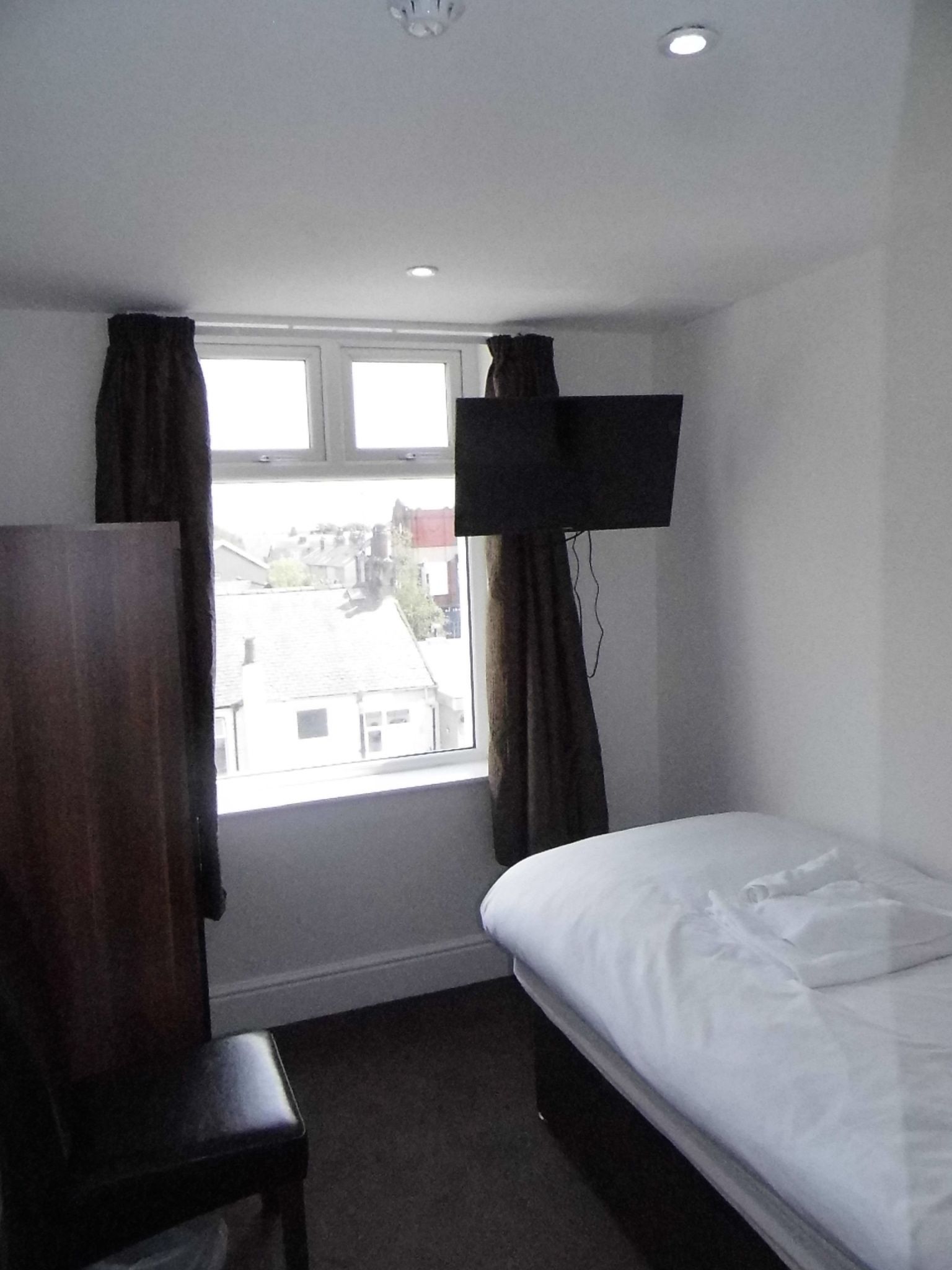 A hotel room with a single bed, a wooden chair, a flat-screen TV mounted on the wall, and a window with curtains.