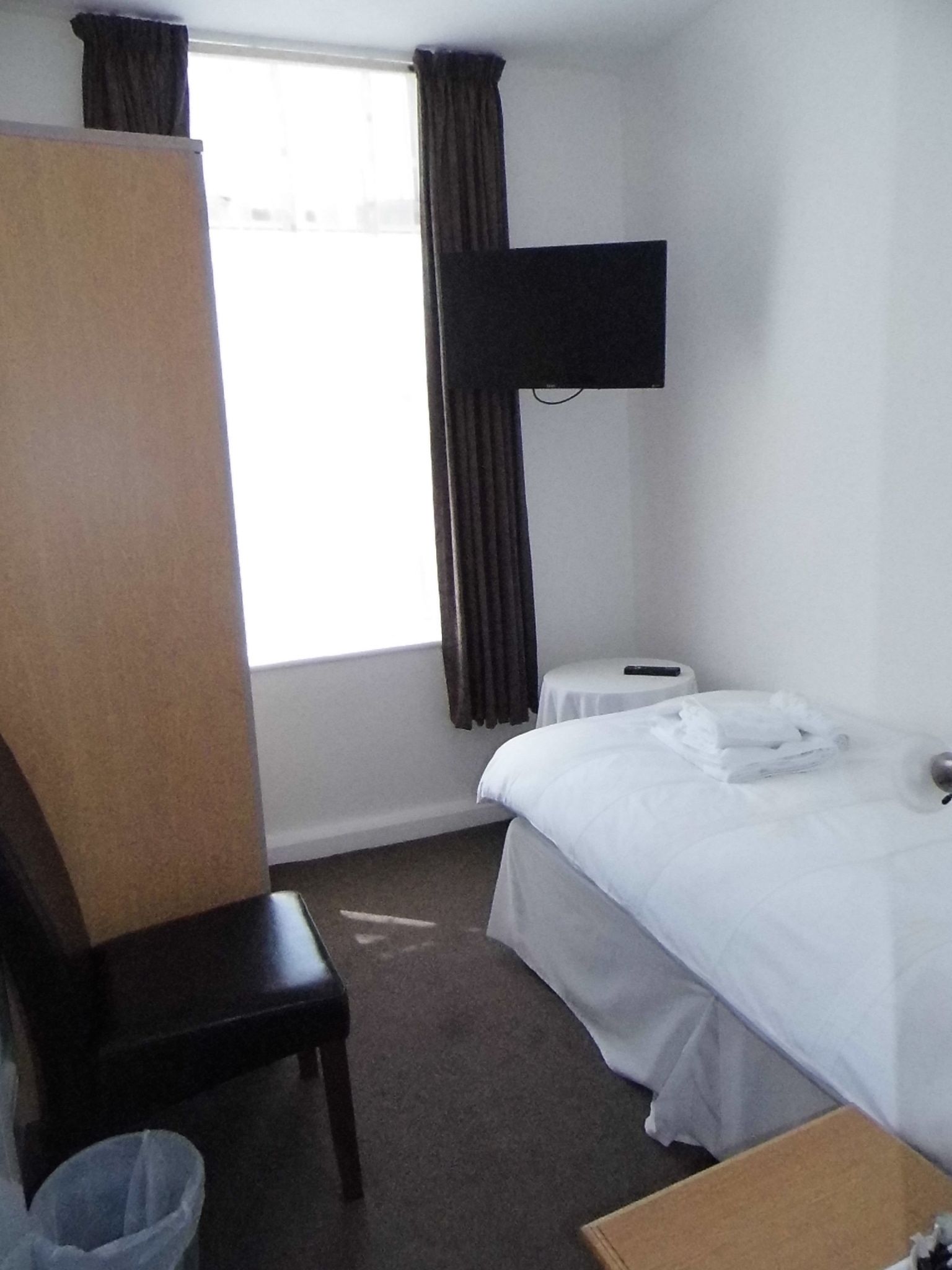 A hotel room with a single bed, a chair, a small table, a trash bin, and a TV mounted on the wall.
