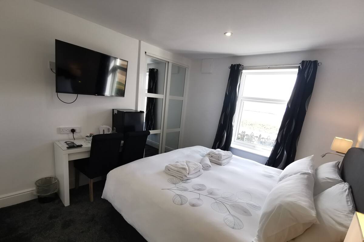 A hotel room with a bed, a desk with a computer, a TV mounted on the wall, and a window with curtains.