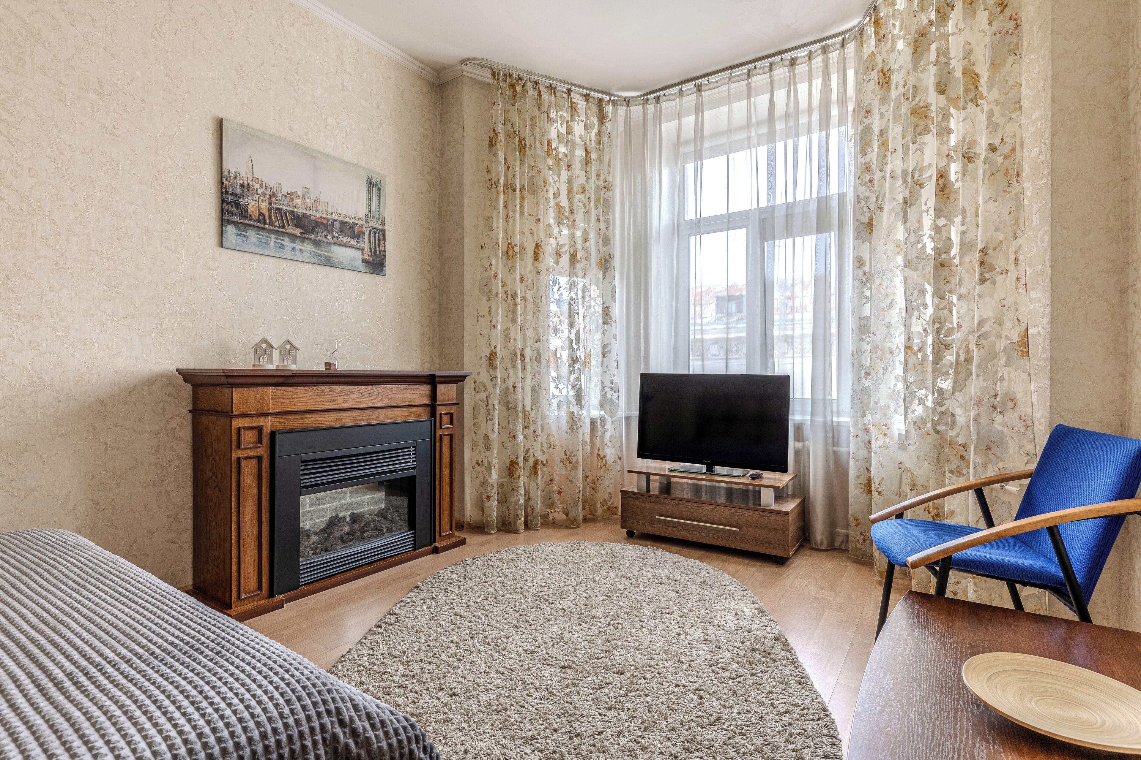 Una sala de estar con chimenea, un televisor de pantalla plana, un sillón azul y un ventanal con cortinas florales.