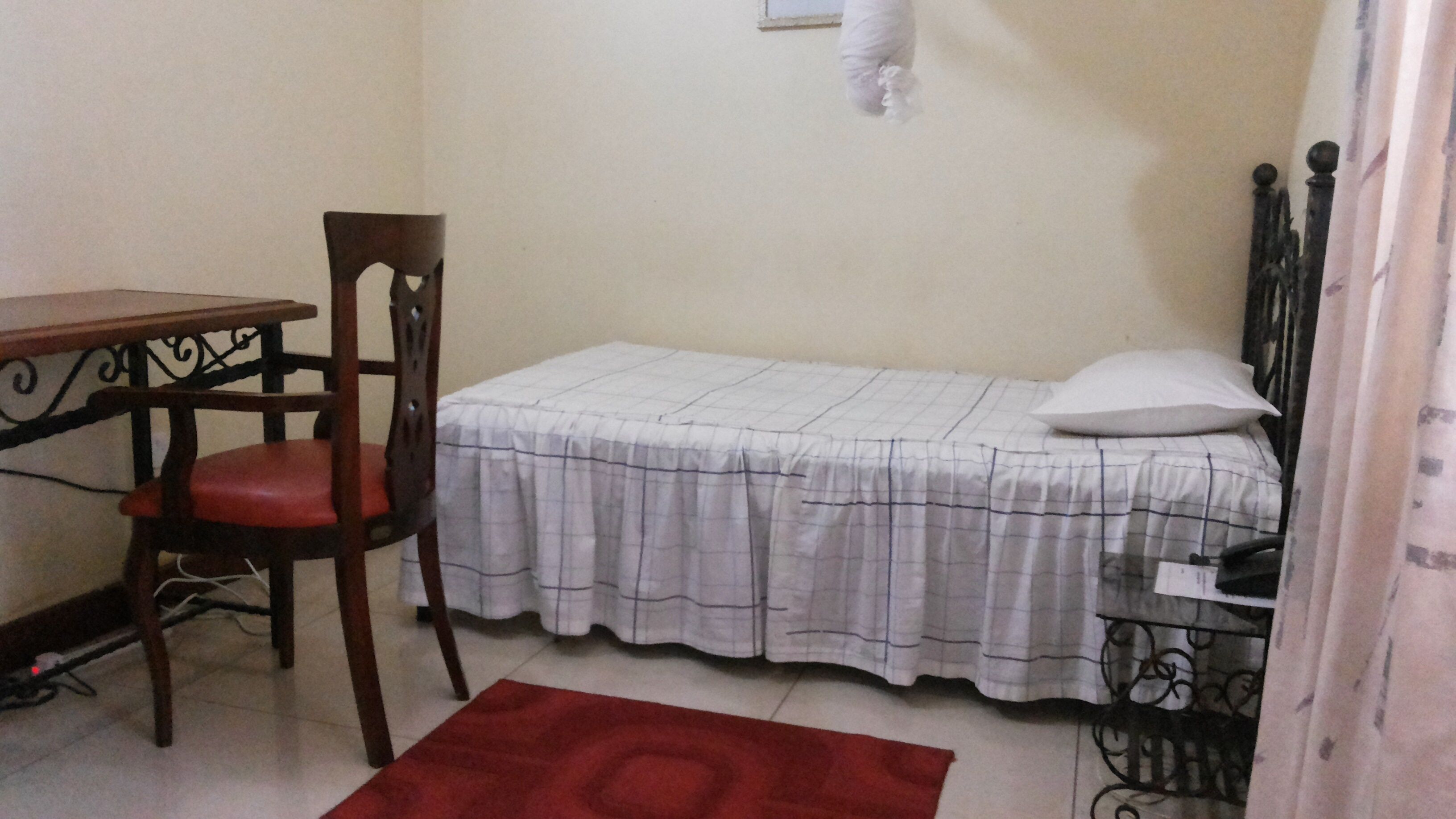 A small room with a single bed, a wooden chair, a table, and a red rug.