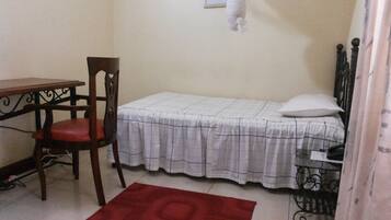 A small room with a single bed, a wooden chair, a table, and a red rug.