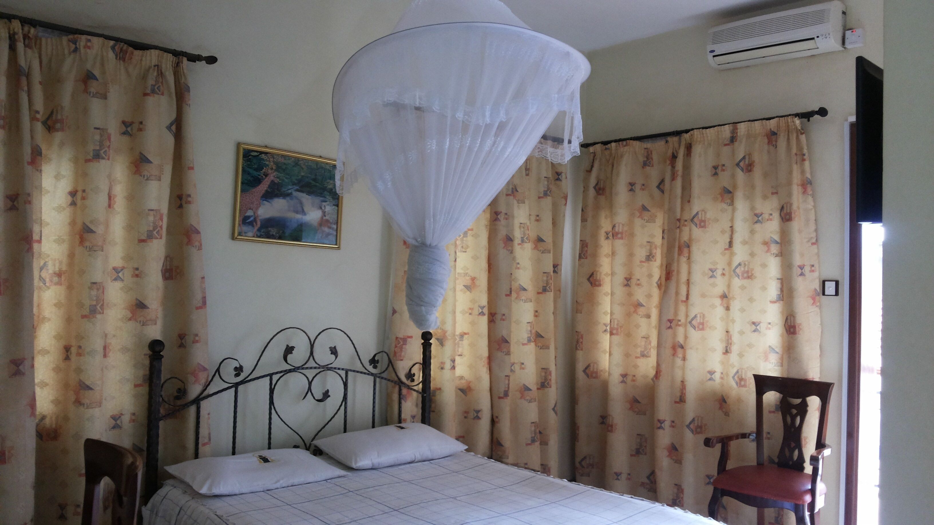 A bedroom with a bed, a chair, curtains, a wall-mounted air conditioner, and a framed picture.