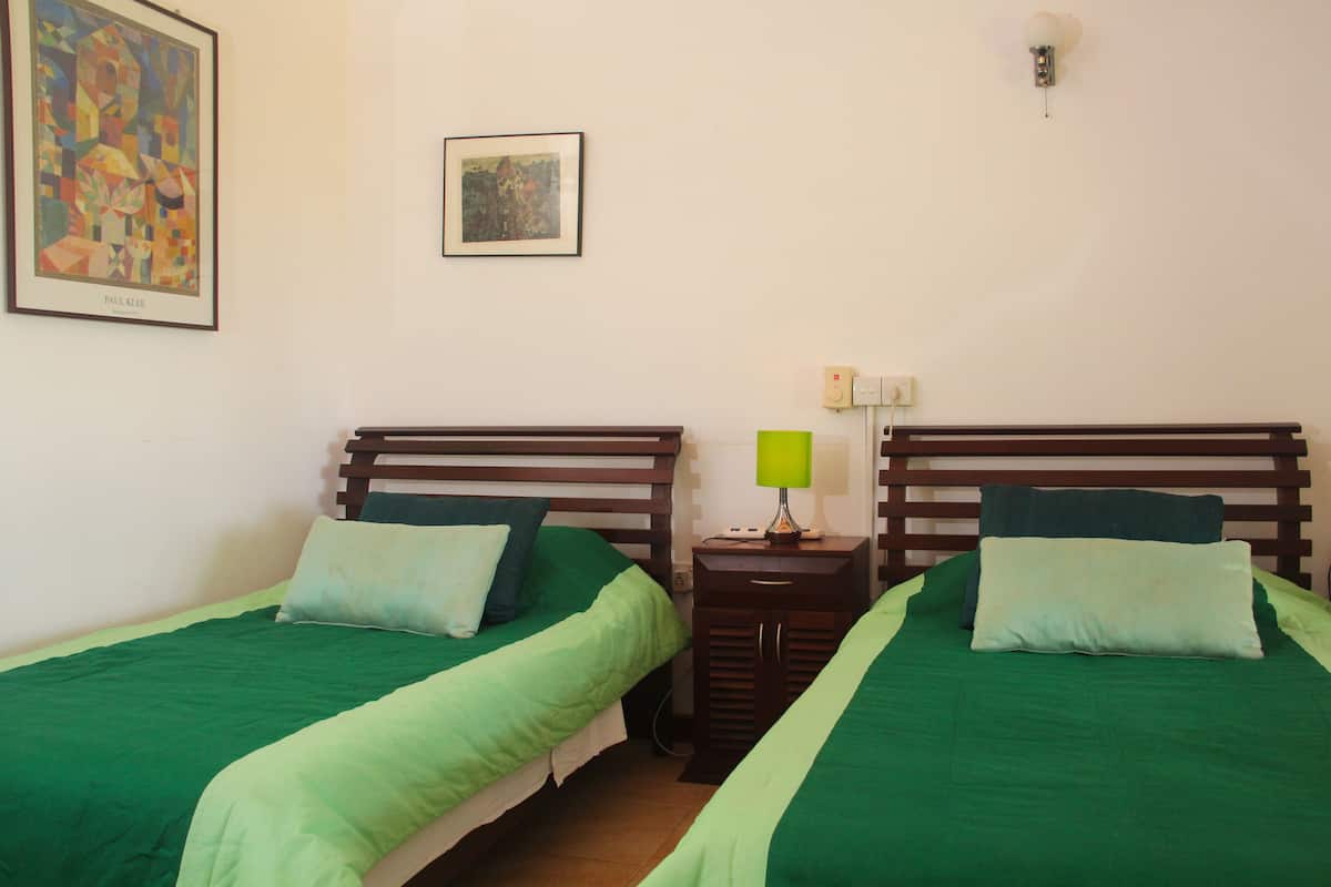 A room with two beds, each with green bedding and pillows, a nightstand with a lamp, and a framed picture on the wall.