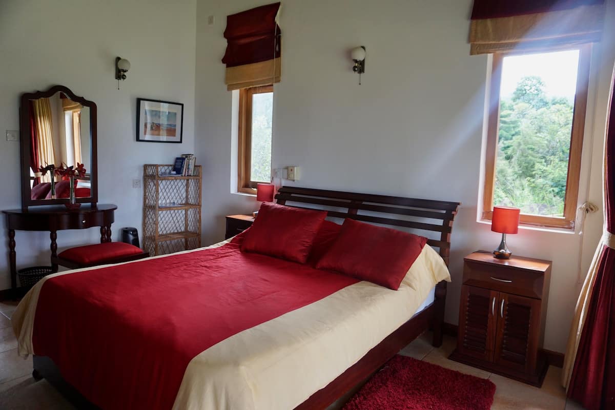 A bedroom with a large bed, a wooden nightstand, a dresser, a mirror, and a window with a view of greenery.