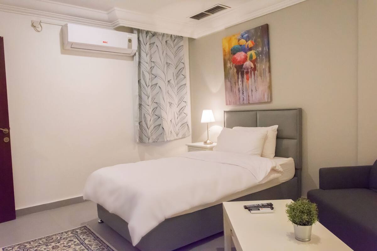 A hotel room with a bed, a nightstand with a lamp, a painting on the wall, and a potted plant on a table.